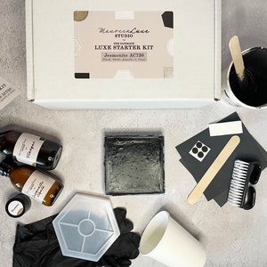 Puede incluir: Una caja blanca y negra con el texto "Maureen Luxe Studio" y "The Ultimate Luxe Starter Kit Jesmonite AC730" en la parte superior. La caja está abierta y el contenido es visible. El contenido incluye una baldosa de resina negra, una taza blanca, un recipiente negro con un palo de madera, dos botellas de líquido, un pequeño recipiente negro, un molde hexagonal blanco, guantes negros, un cepillo negro, un trozo de papel negro con puntos blancos y un palo de madera.