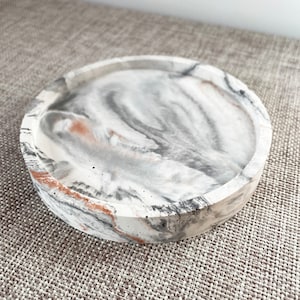 Pu&ograve; includere: Un vassoio rotondo in cemento bianco con un design marmoreo in tonalit&agrave; di grigio e marrone. Il vassoio ha un bordo rialzato ed &egrave; perfetto per contenere piccoli oggetti.