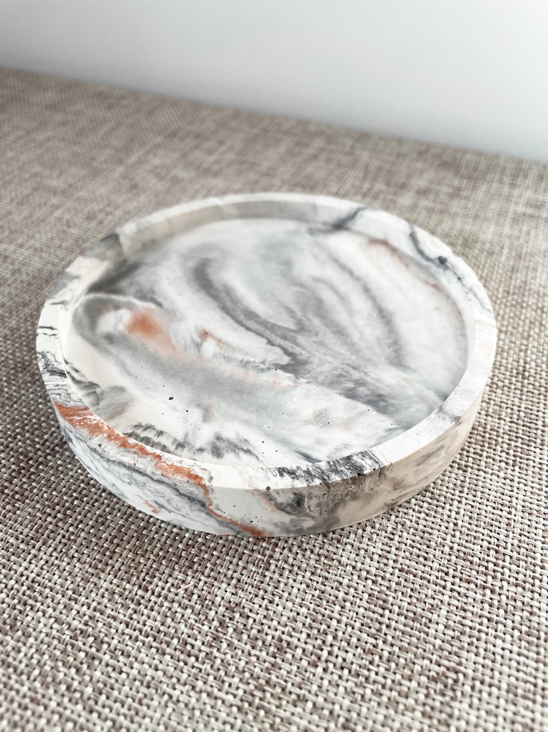 Pu&ograve; includere: Un vassoio rotondo in cemento bianco con un design marmoreo in tonalit&agrave; di grigio e marrone. Il vassoio ha un bordo rialzato ed &egrave; perfetto per contenere piccoli oggetti.