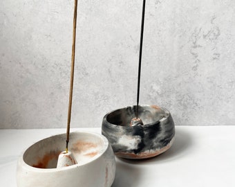 May include: Two white ceramic incense holders with a marbled design. One holder has a black and white marbled design and the other has a light brown and white marbled design. Both holders have a small cone in the center for incense sticks.