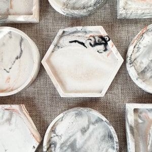 Pu&ograve; includere: Un set di sette vassoi decorativi in ​​calcestruzzo con un design marmoreo in tonalit&agrave; di bianco, grigio e marrone. I vassoi sono di varie forme, tra cui rotonda, quadrata, esagonale e rettangolare.