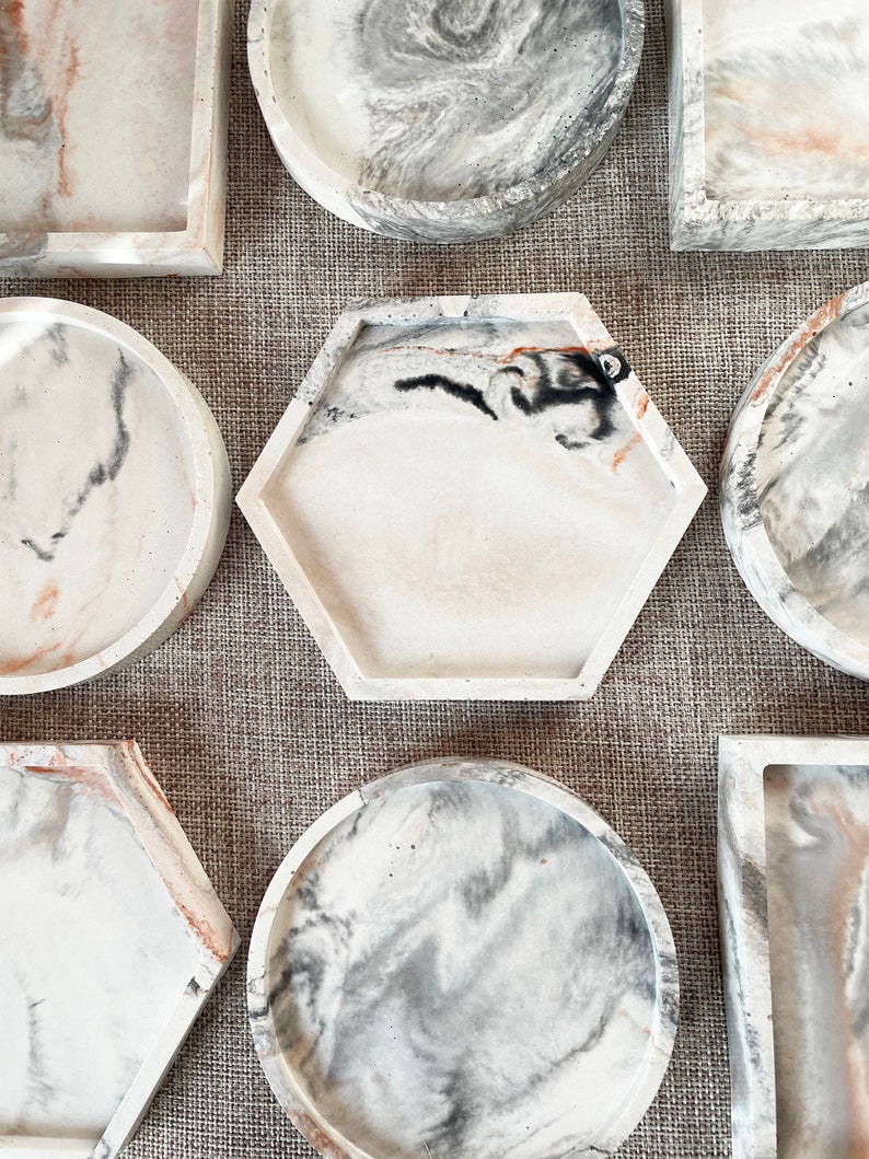 Pu&ograve; includere: Un set di sette vassoi decorativi in ​​calcestruzzo con un design marmoreo in tonalit&agrave; di bianco, grigio e marrone. I vassoi sono di varie forme, tra cui rotonda, quadrata, esagonale e rettangolare.