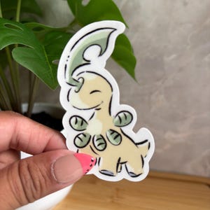 May include: A die-cut vinyl sticker featuring a cartoon creature with a pale yellow body, a curved green leaf-like appendage on its head, and small green circular markings on its chest, outlined in a thick white border against a plain background.