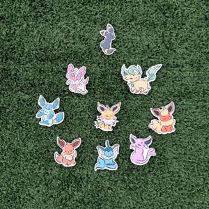 May include: A collection of colorful cartoon character stickers, each with a unique design. The stickers feature various creatures in shades of blue, pink, yellow, orange, and green, with black outlines. The stickers are arranged on a green surface.