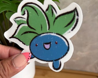 May include: A die-cut sticker featuring a cartoon character with a round blue body and green leaves sprouting from the top. The character has a smiling face with a purple mouth, small red eyes, and a textured, hand-drawn black outline style.
