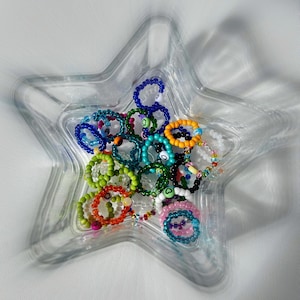 May include: A collection of colorful beaded rings in a clear star-shaped dish. The rings are made of various colors, including blue, green, yellow, and red.