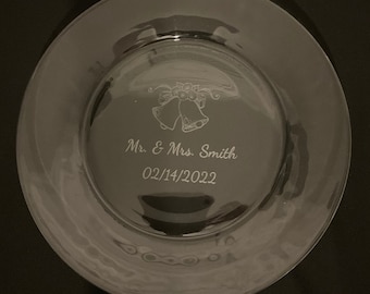 May include: A clear glass plate with an engraved design of two wedding bells and a floral wreath. The text "Mr. & Mrs. Smith 02/14/2022" is engraved below the bells.