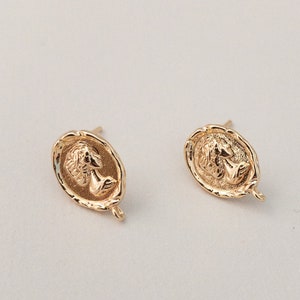 May include: A pair of gold-toned earrings with a detailed design of a woman's face. The earrings are oval-shaped and have a textured surface.