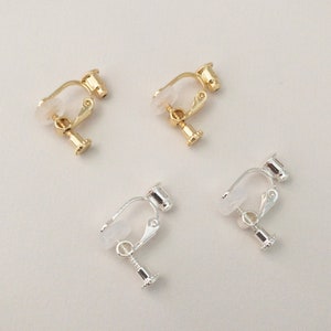 May include: Four clip-on earrings in gold and silver. Each earring features a small, bell-shaped charm suspended from a clip. The clip-on mechanism is visible, with a clear, plastic pad for comfort. The earrings are presented against a light background.