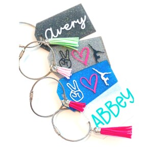 May include: Four colorful luggage tags with silver key rings and tassels. The tags feature glitter, peace signs, hearts, and the names "Avery" and "Abbey".