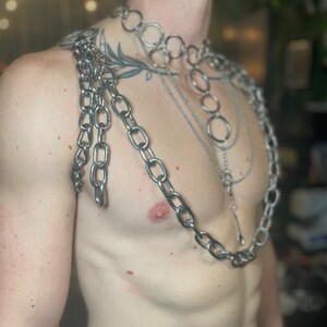 Op de afbeelding: Een persoon draagt meerdere zilveren kettingen om hun nek en schouders. De kettingen zijn gelaagd en omvatten een dikke ketting met grote schakels, een dunnere ketting met kleinere schakels en een ketting met ronde schakels.