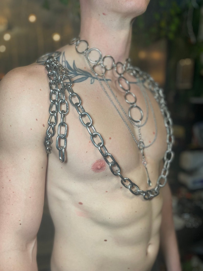 Op de afbeelding: Een persoon draagt meerdere zilveren kettingen om hun nek en schouders. De kettingen zijn gelaagd en omvatten een dikke ketting met grote schakels, een dunnere ketting met kleinere schakels en een ketting met ronde schakels.