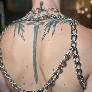 Op de afbeelding: Een zilveren ketting met een groot, dik kettinglinkontwerp. De ketting wordt om de nek en schouders gedragen en wordt over de rug van de drager gelegd. De ketting is gemaakt van metaal en heeft een glanzende afwerking.