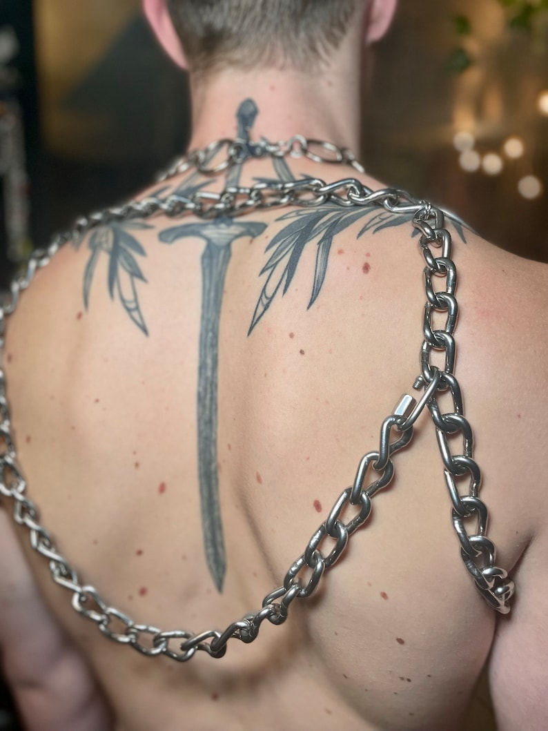 Op de afbeelding: Een zilveren ketting met een groot, dik kettinglinkontwerp. De ketting wordt om de nek en schouders gedragen en wordt over de rug van de drager gelegd. De ketting is gemaakt van metaal en heeft een glanzende afwerking.