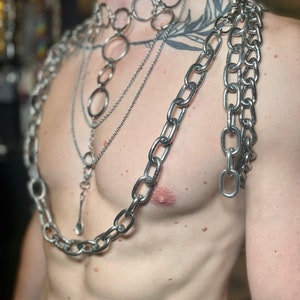 Op de afbeelding: Een persoon draagt meerdere zilveren kettingen om de nek en schouders. De kettingen zijn van verschillende diktes en stijlen, waaronder een dikke ketting met grote schakels, een dunnere ketting met kleinere schakels en een ketting met ronde schakels. De kettingen zijn in lagen gerangschikt, met de dikste ketting onderaan en de dunste ketting bovenaan.