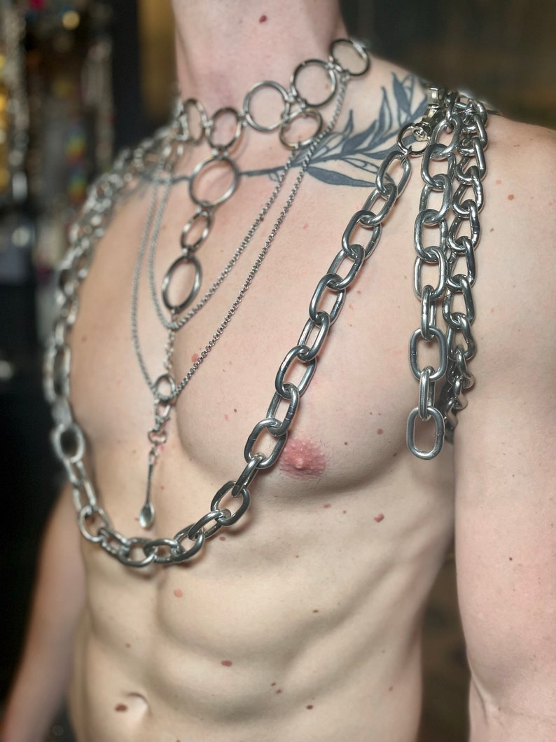 Op de afbeelding: Een persoon draagt meerdere zilveren kettingen om de nek en schouders. De kettingen zijn van verschillende diktes en stijlen, waaronder een dikke ketting met grote schakels, een dunnere ketting met kleinere schakels en een ketting met ronde schakels. De kettingen zijn in lagen gerangschikt, met de dikste ketting onderaan en de dunste ketting bovenaan.