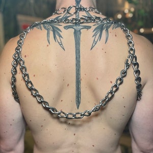 Op de afbeelding: Een persoon met een grote zwaardtattoo op zijn rug, die een zilveren kettingketting en twee zilveren kettingen om zijn schouders draagt.