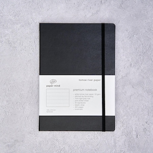 May include: A black hardcover notebook with a vertical black elastic strap. A white paper band wraps around the centre, featuring the text paper mind, tomoe river paper, premium notebook, and specifications including 148 x 210 mm dimensions.