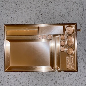 May include: A gold-colored rolling tray with multiple compartments and a built-in grinder. The tray has a smooth, metallic finish and features a raised design.