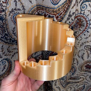 May include: A 3D printed, beige-colored, wall-mounted organizer with multiple holes for storing items. The organizer has a rounded shape and a flat back.