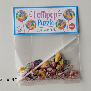 May include: A clear plastic bag containing a 3-D lollipop puzzle with 2584 pieces. The bag has a label that says "3-D Lollipop Puzzle" and "2584+ PIECES". The label also has a picture of a lollipop and the words "New!" and "3y+".
