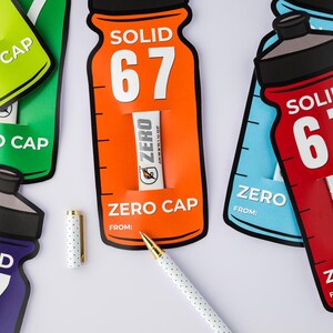 May include: A collection of bottle-shaped cards in green, orange, blue, and red, each displaying "SOLID 67" and "ZERO CAP". Small white tubes with the "ZERO" logo are visible. A white pen with gold details and a dotted cap are also present.