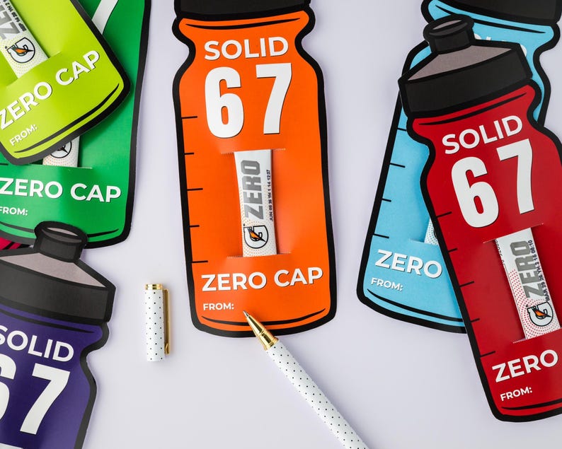 May include: A collection of bottle-shaped cards in green, orange, blue, and red, each displaying "SOLID 67" and "ZERO CAP". Small white tubes with the "ZERO" logo are visible. A white pen with gold details and a dotted cap are also present.