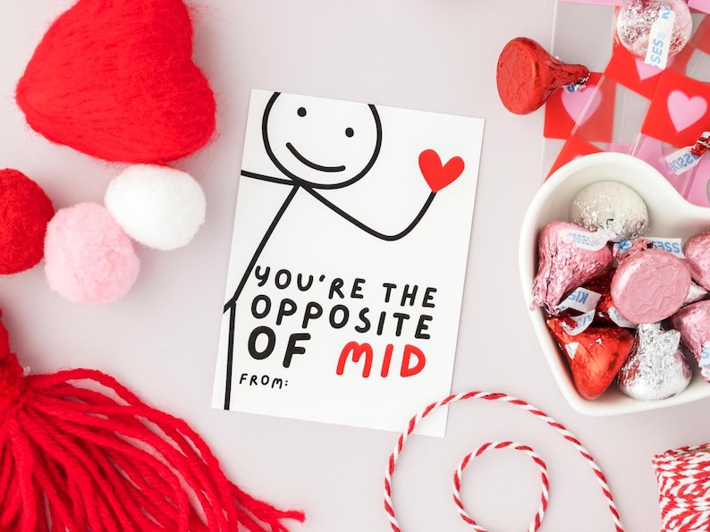 Op de afbeelding: Een witte kaart met zwarte tekst die "You're the opposite of mid" zegt met een rood hart getekend naast de tekst. De kaart heeft een stokfiguurtekening met een rood hart in zijn hand. De kaart is omringd door rode harten, witte pompoenen en rood en wit touw.