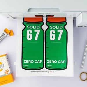 May include: A printed sheet featuring two green bottle designs with orange tops, the word "SOLID" and the number "67". Below is "ZERO CAP". Also visible are orange drink mix sachets, a box of orange drink mix, a pen, and gold scissors.