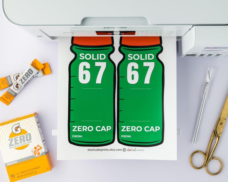 May include: A printed sheet featuring two green bottle designs with orange tops, the word "SOLID" and the number "67". Below is "ZERO CAP". Also visible are orange drink mix sachets, a box of orange drink mix, a pen, and gold scissors.