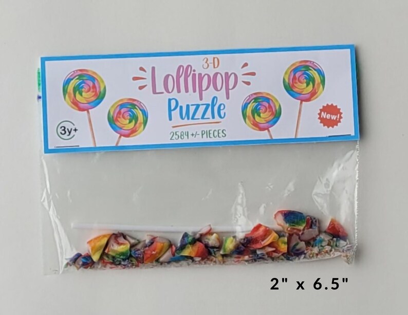 May include: A plastic bag containing a 3-D lollipop puzzle with 2584+ pieces. The bag has a colorful label that says "3-D Lollipop Puzzle" and "2584+ Pieces". The label also has a picture of two lollipops and the words "New!" and "3y+". The puzzle pieces are scattered inside the bag. The bag is 2 inches by 6.5 inches.
