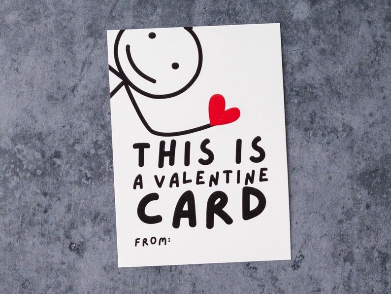 May include: A white Valentine's Day card with a stick figure holding a red heart. The card reads "THIS IS A VALENTINE CARD" in bold black letters, with a space for "FROM:" at the bottom. The background is a textured gray surface.