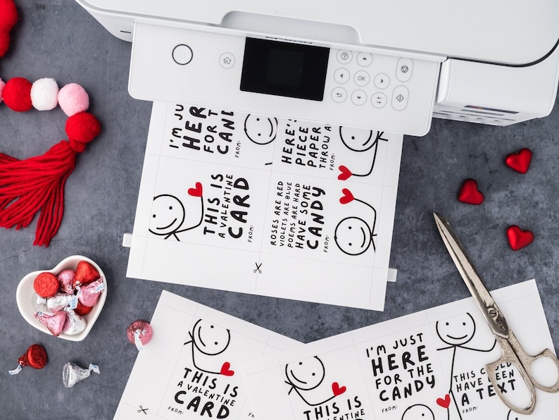 May include: A white printer printing Valentine's Day cards with stick figure illustrations and text. The cards have phrases like "I'm just here for the candy" and "This is a Valentine card." Red heart-shaped candies and a pair of scissors are also visible.