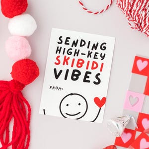Op de afbeelding: Een witte kaart met zwarte en rode tekst die "Sending High-Key Skibidi Vibes" zegt met een smiley-tekening en een rood hart. De kaart is omringd door rode en witte pompoenen, rood en wit touw en een roze en rood geruit patroon.