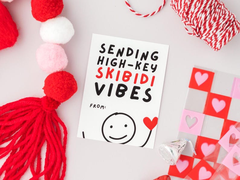 Op de afbeelding: Een witte kaart met zwarte en rode tekst die "Sending High-Key Skibidi Vibes" zegt met een smiley-tekening en een rood hart. De kaart is omringd door rode en witte pompoenen, rood en wit touw en een roze en rood geruit patroon.