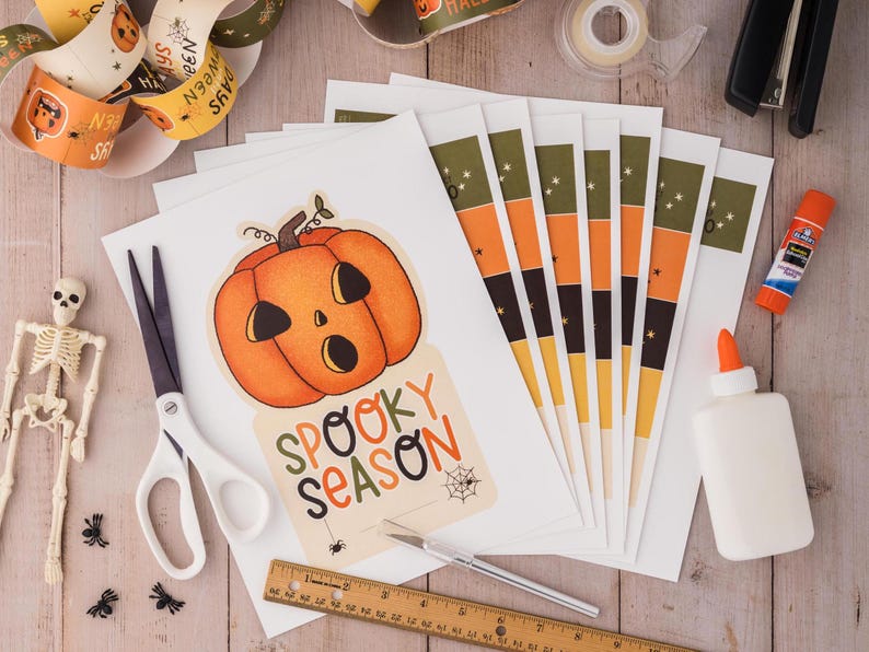 May include: A craft project with a printable Halloween card featuring a cartoon pumpkin with the text "Spooky Season". The image also shows craft supplies such as scissors, glue, and a ruler.