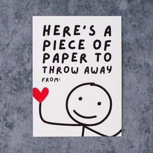 May include: A white card with black text that reads "HERE'S A PIECE OF PAPER TO THROW AWAY" and "FROM:". A stick figure holds a red heart. The card is on a gray surface.
