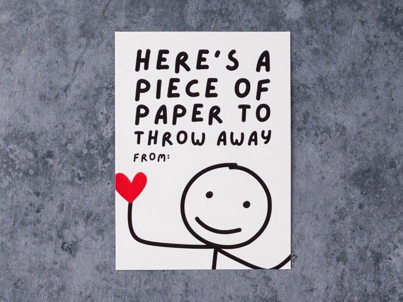 May include: A white card with black text that reads "HERE'S A PIECE OF PAPER TO THROW AWAY" and "FROM:". A stick figure holds a red heart. The card is on a gray surface.