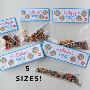 May include: Five different sizes of clear plastic bags filled with colorful candy pieces and a lollipop stick. Each bag has a label that reads "3-D Lollipop Puzzle 2581+ Pieces" with a rainbow lollipop graphic. The label also says "New!" and "3y+".