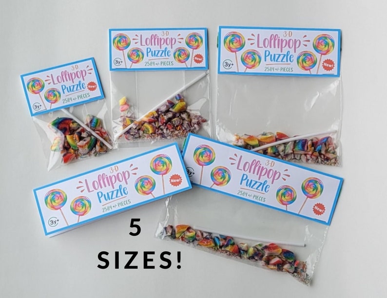 May include: Five different sizes of clear plastic bags filled with colorful candy pieces and a lollipop stick. Each bag has a label that reads "3-D Lollipop Puzzle 2581+ Pieces" with a rainbow lollipop graphic. The label also says "New!" and "3y+".