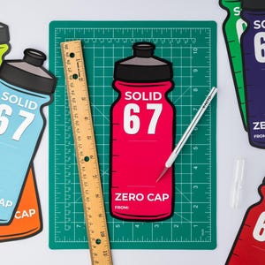 May include: A selection of brightly coloured water bottle cutouts, each displaying "SOLID 67" and "ZERO CAP". A wooden ruler and a craft knife rest on a green cutting mat. The bottles are in red, blue, orange, green, and purple.