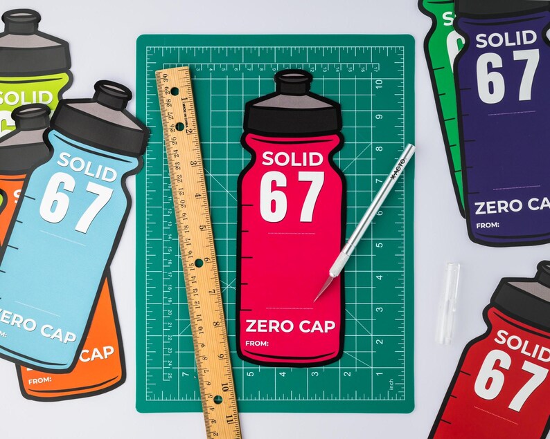 May include: A selection of brightly coloured water bottle cutouts, each displaying "SOLID 67" and "ZERO CAP". A wooden ruler and a craft knife rest on a green cutting mat. The bottles are in red, blue, orange, green, and purple.