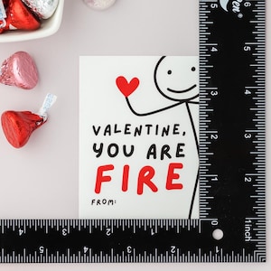 Op de afbeelding: Een witte kaart met een rood hart en een stick figure-illustratie. De kaart luidt "Valentine, you are fire" met het woord "fire" in rood. De kaart heeft ook de woorden "from:" onderaan.