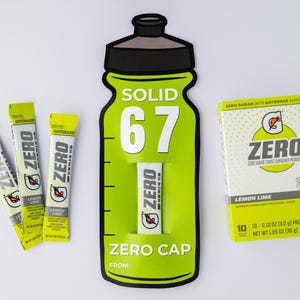 May include: A still-life image featuring Gatorade Zero products. The composition includes a large green bottle-shaped display with the text "SOLID 67" and "ZERO CAP", along with individual packets and a box of Lemon Lime flavoured powder. The packets and box feature the "ZERO" logo.