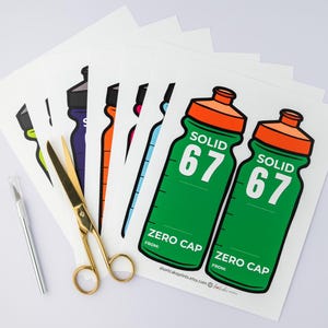 May include: A selection of water bottle stickers in different colours, including green, orange, purple, and blue. The stickers display the text "SOLID 67" and "ZERO CAP". Gold scissors and a silver craft knife are also visible.
