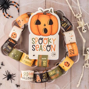 May include: A Halloween countdown chain with orange and black paper rings featuring numbers and the text "Days until Halloween". A large orange pumpkin with a spooky face and the text "Spooky Season" is in the center. Two white skeletons are on either side of the chain.