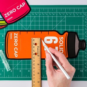 May include: An orange and black Zero Cap bottle-shaped cutout is being measured with a ruler and a craft knife on a green cutting mat. The cutout has the text "ZERO CAP" and "FROM:" printed on it. A clear plastic ruler and other cutouts are also visible.