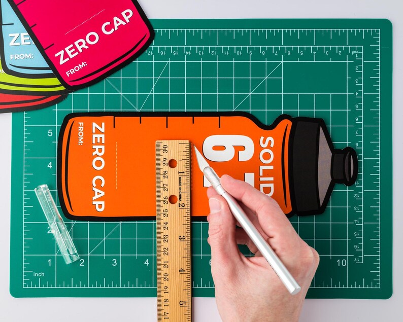 May include: An orange and black Zero Cap bottle-shaped cutout is being measured with a ruler and a craft knife on a green cutting mat. The cutout has the text "ZERO CAP" and "FROM:" printed on it. A clear plastic ruler and other cutouts are also visible.