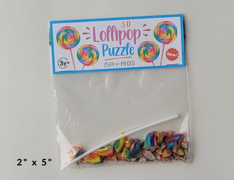 May include: A clear plastic bag containing a 3-D lollipop puzzle with 2584+ pieces. The bag has a colorful label that says "3-D Lollipop Puzzle" and "2584+ Pieces". The label also has a picture of a lollipop and the words "New!" and "3y+".