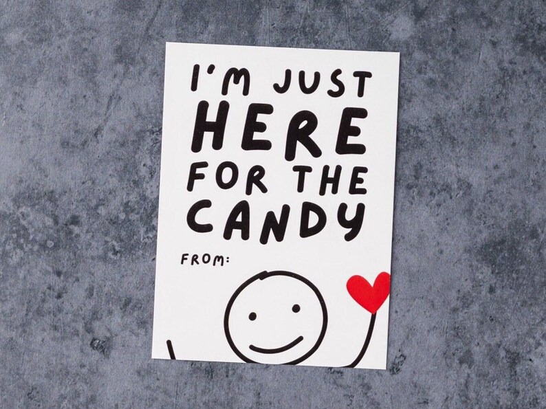 May include: A white card with the text "I'M JUST HERE FOR THE CANDY" in black, bold letters. A stick figure with a smiling face and a red heart is drawn below the text. The card is on a gray surface.